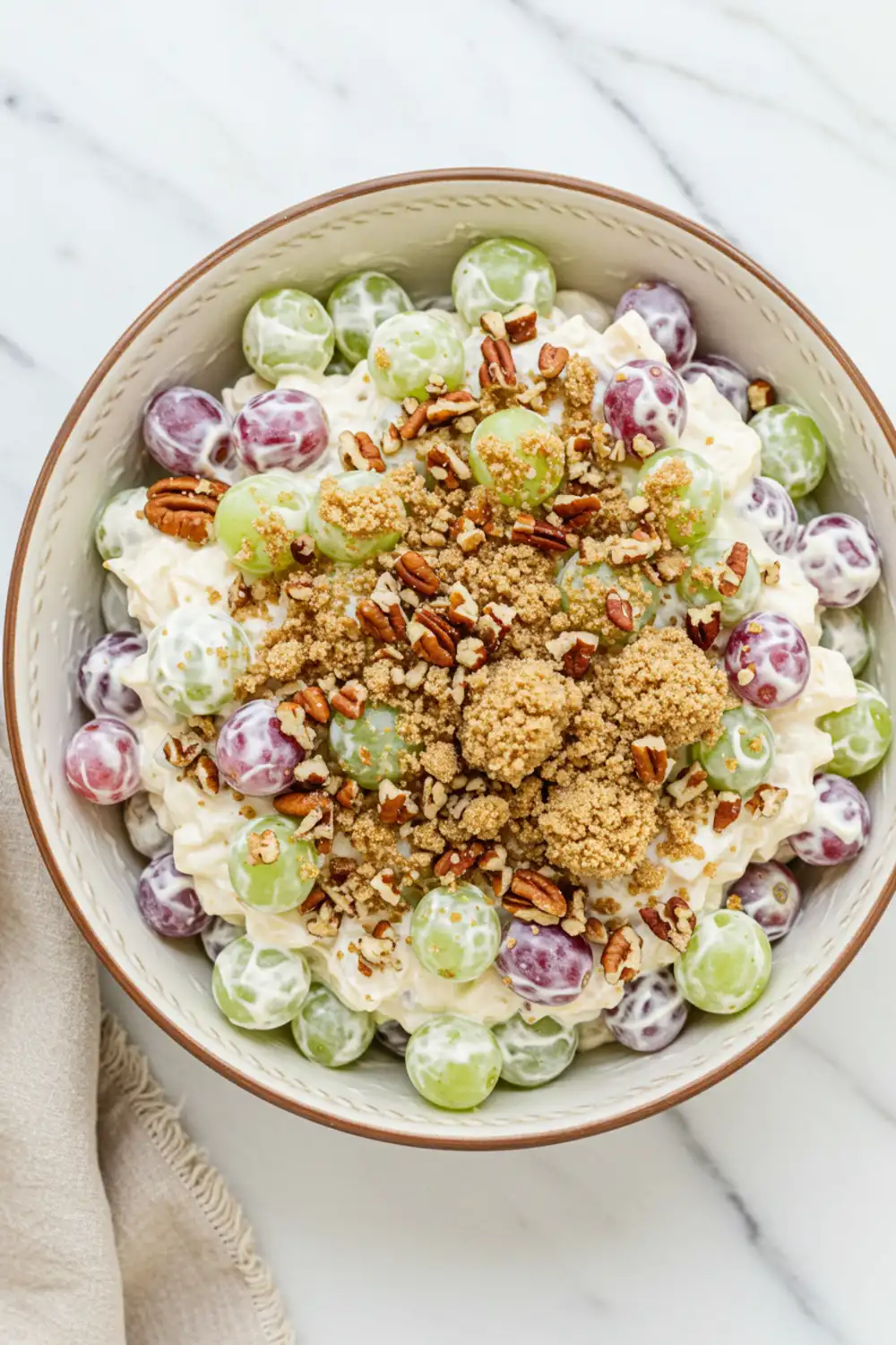 Close-up top-down view of the finished Chicken Salad Chick Grape Salad in a serving bowl, showing creamy coated green and red grapes generously topped with a crunchy brown sugar-pecan mixture, looking natural and home-prepared.
