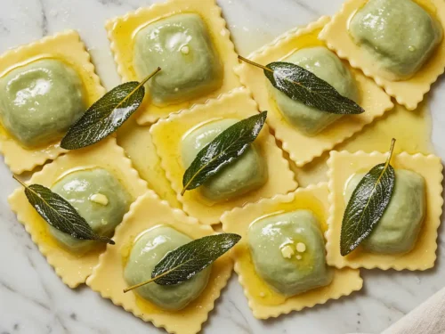 Flat-lay close-up of spinach ricotta ravioli tossed in glistening nutty golden brown butter sauce with naturally crisped fresh sage leaves, presented on a light marble background with a crumpled beige cloth in natural light.