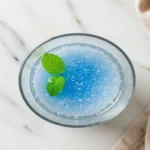 Close-up top-down view of the finished Blue Salt Trick Recipe elixir in a clear glass, showing clear water with a subtle shimmer, optional mint garnish, looking natural and refreshing on a light marble surface.
