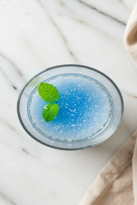 Close-up top-down view of the finished Blue Salt Trick Recipe elixir in a clear glass, showing clear water with a subtle shimmer, optional mint garnish, looking natural and refreshing on a light marble surface.