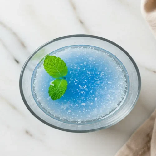 Close-up top-down view of the finished Blue Salt Trick Recipe elixir in a clear glass, showing clear water with a subtle shimmer, optional mint garnish, looking natural and refreshing on a light marble surface.