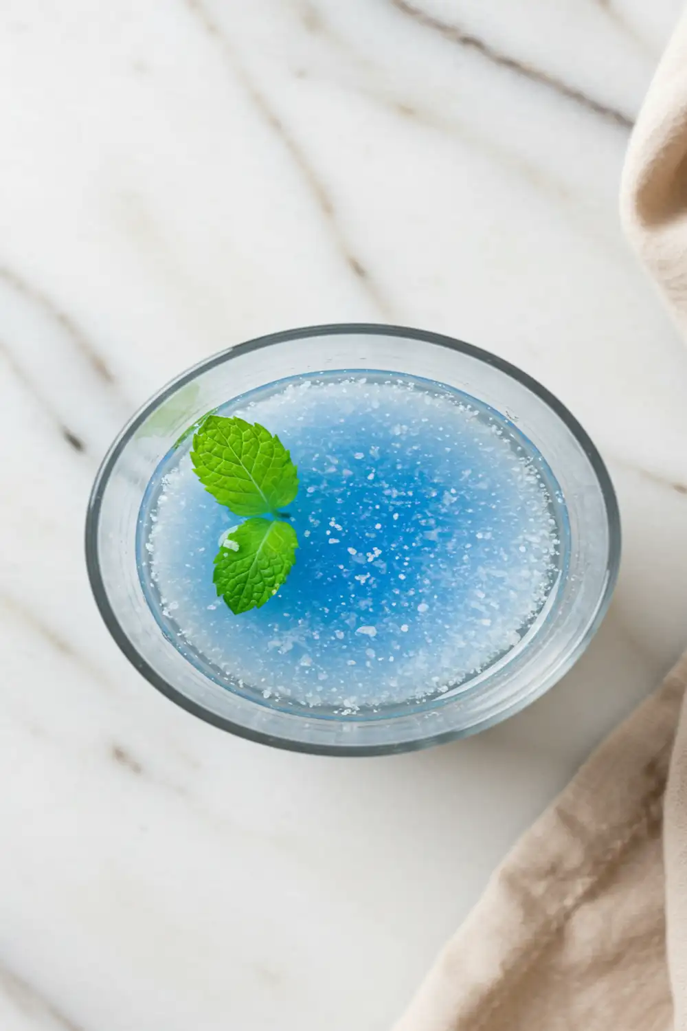 Close-up top-down view of the finished Blue Salt Trick Recipe elixir in a clear glass, showing clear water with a subtle shimmer, optional mint garnish, looking natural and refreshing on a light marble surface.
