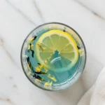 Close-up top-down view of the finished Brazilian Mounjaro inspired wellness drink in a clear glass, showing suspended ginger and its final color, looking natural, refreshing, and home-prepared on a light marble surface.