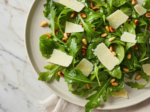 Overhead close-up of a peppery arugula salad with shaved parmesan and toasted pine nuts, lightly dressed, displayed naturally on a light marble surface with a beige cloth under soft daylight.