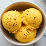 Extreme close-up overhead view of creamy, vibrant orange spicy mango ice cream scoops in a bowl, showing visible reddish spice specks throughout and a chili lime seasoning garnish on top.