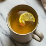 Close-up top-down view of a hand holding a sturdy mug of Pink Salt and Turmeric Morning Tonic garnished with lemon, set on a natural surface with a journal, looking natural and part of a morning ritual.